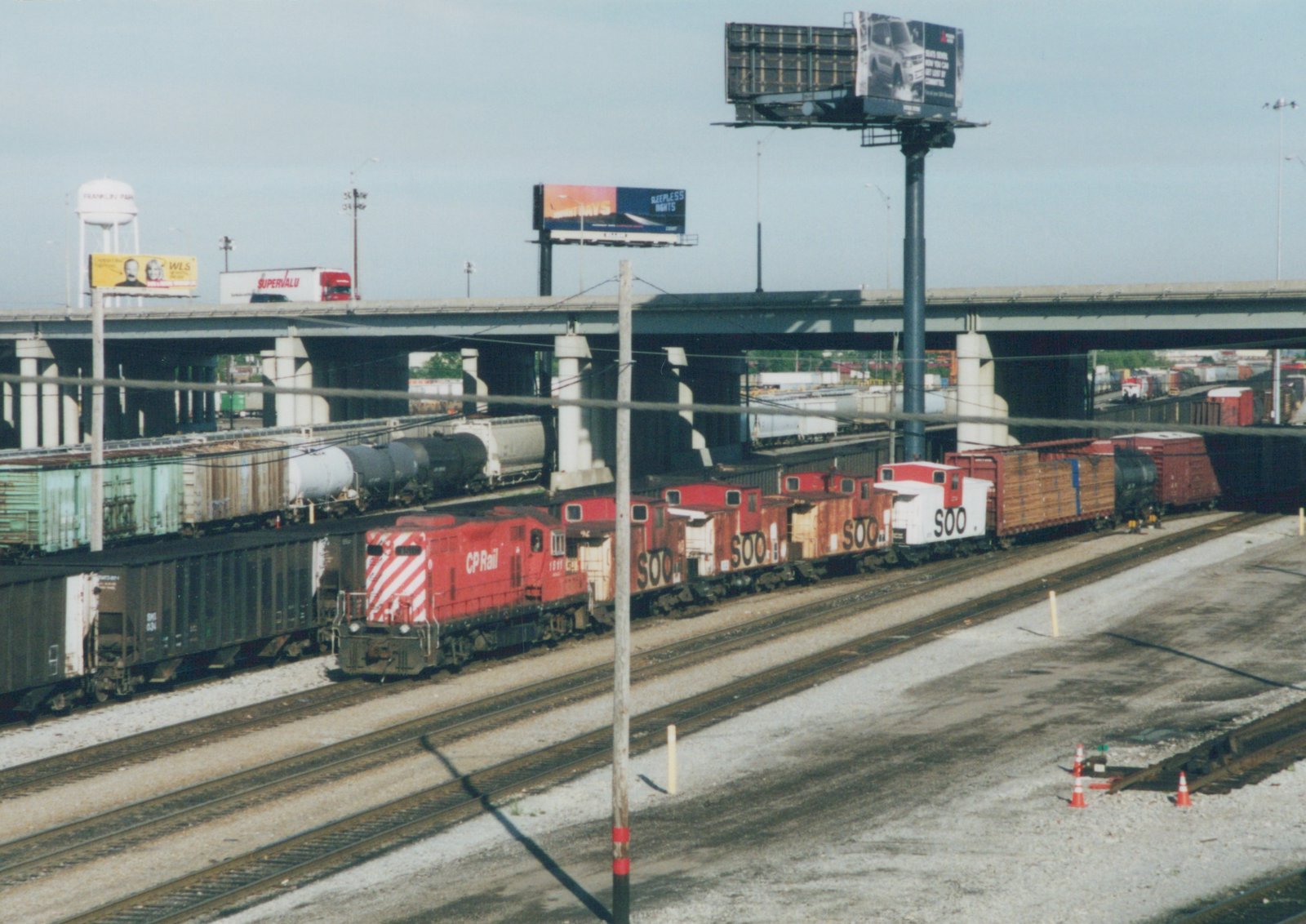 A red CP Rail locomotive with the number 1597 is pulling a train with several cabooses marked 'SOO' under an elevated highway, with a water tower and billboards in the background.