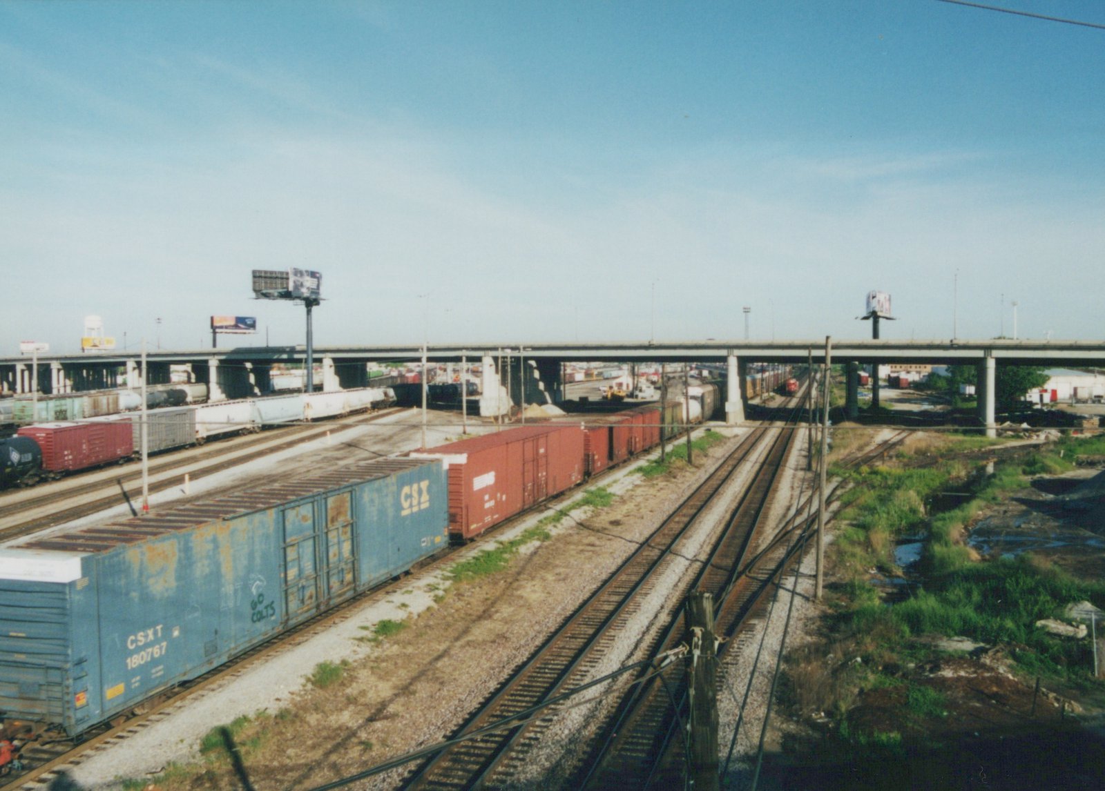 A blue CSX freight car with the number 180767 and 'GO COLTS' graffiti is parked on a track in a rail yard under a large concrete highway bridge on a clear day.