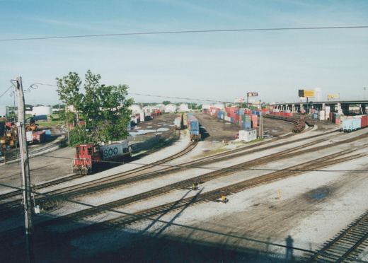 A red and white SOO Line diesel locomotive, number 35, sits on tracks in a large rail yard filled with shipping containers, viewed from an elevated position under a clear blue sky.