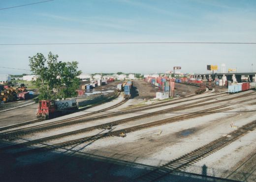 An elevated view of a Soo Line SW1200 locomotive moving through a sprawling rail yard filled with freight cars and shipping containers, located in Franklin Park, Illinois.