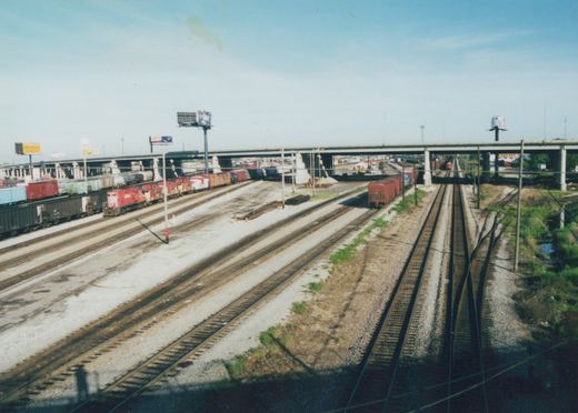 An elevated view of a sprawling railroad yard in Franklin Park under a large concrete highway bridge, with multiple tracks and a red locomotive visible.