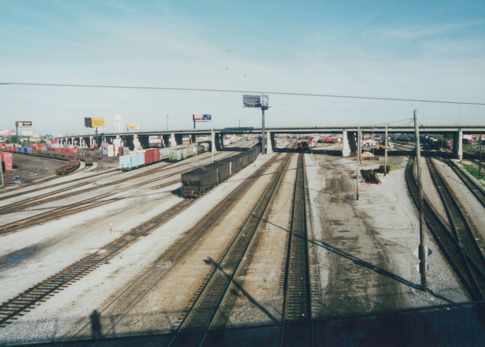 A high-angle view of a sprawling railroad yard with many tracks, freight cars, and a concrete overpass in the background under a clear blue sky.
