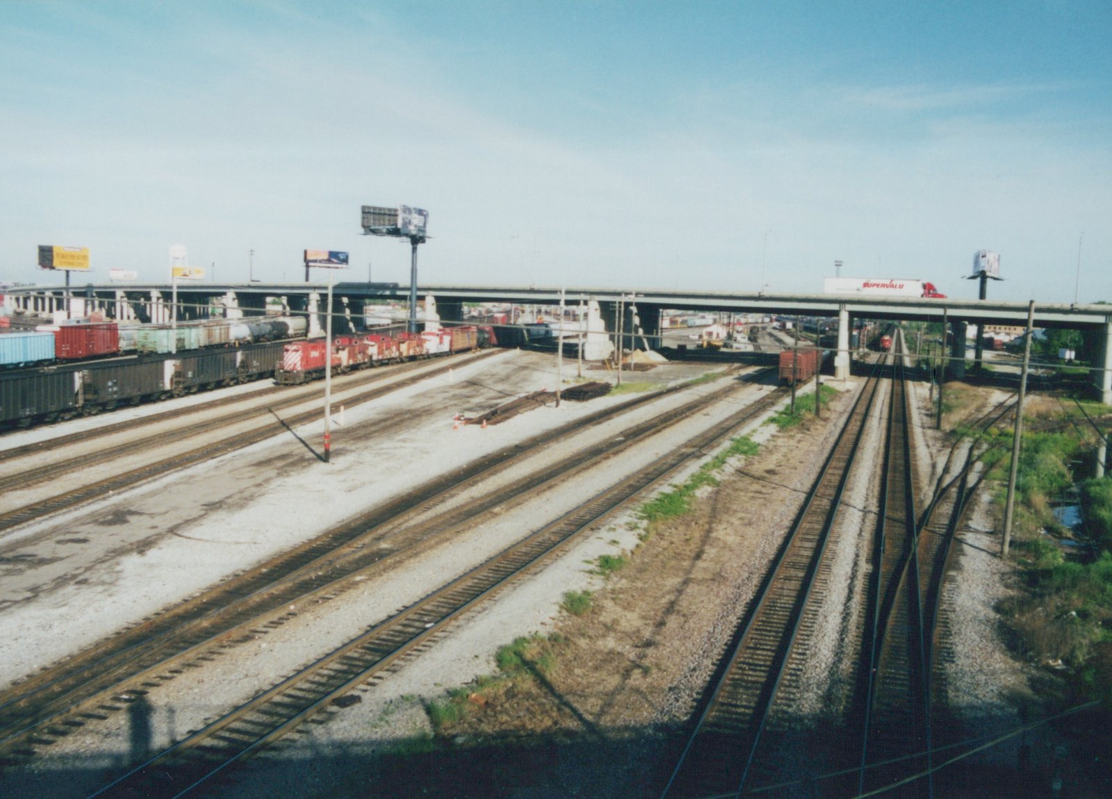 An overhead view of a Soo Line diesel locomotive pulling a freight train through a yard with multiple tracks, under a concrete bridge carrying a semi-trailer with 'SUPERVALU' lettering.