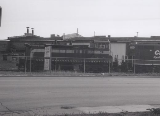 A black and white photo of a Canadian Pacific locomotive with the number 8254, behind a chain-link fence, with a sign reading 'CP Rail System Bensenville Diesel House' nearby.