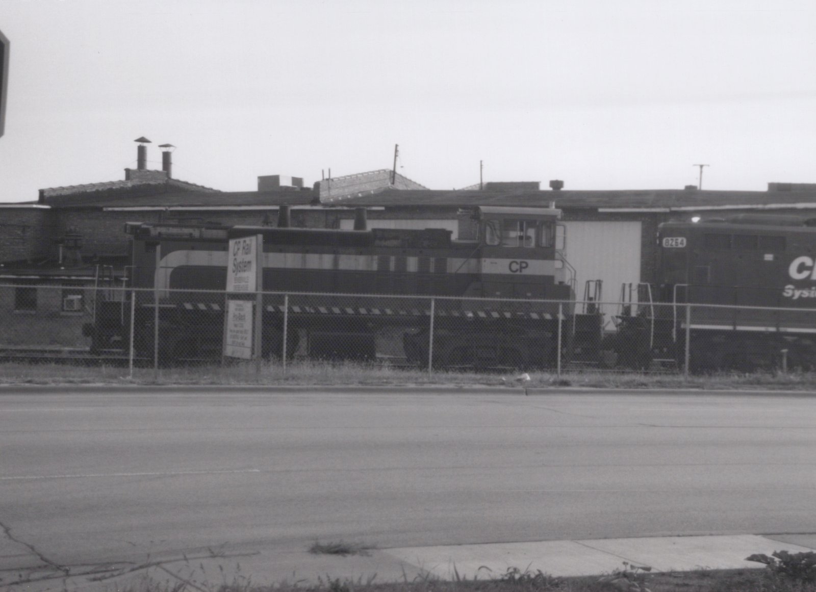 A black and white photo of a Canadian Pacific locomotive with the number 8254, behind a chain-link fence, with a sign reading 'CP Rail System Bensenville Diesel House' nearby.