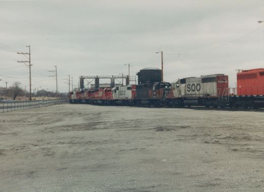 Side view of several Soo Line locomotives on a track in a gravel yard, under a grey, overcast sky. The engines are painted in red, white, and black livery with 'SOO' branding.