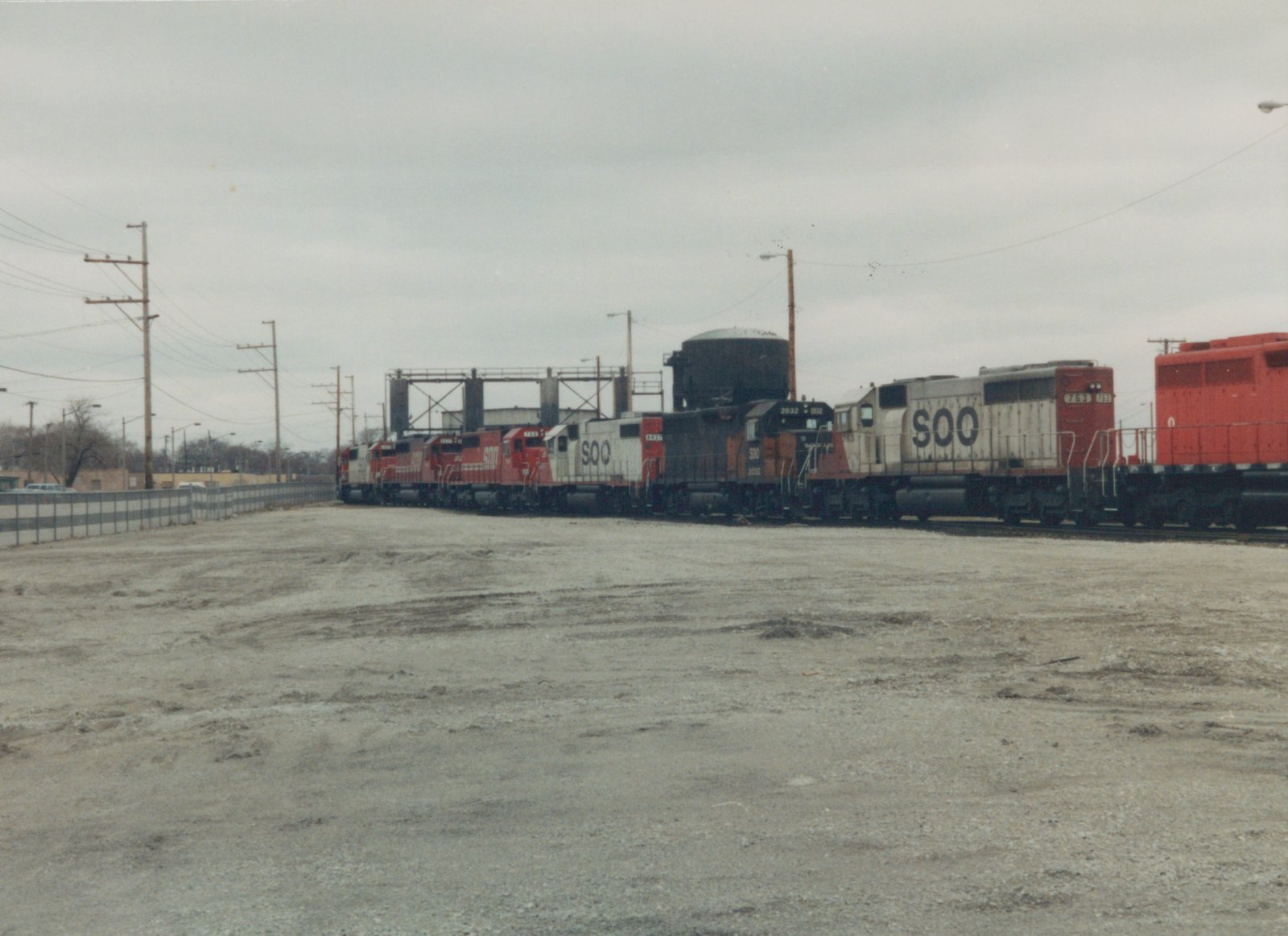 Side view of several Soo Line locomotives on a track in a gravel yard, under a grey, overcast sky. The engines are painted in red, white, and black livery with 'SOO' branding.