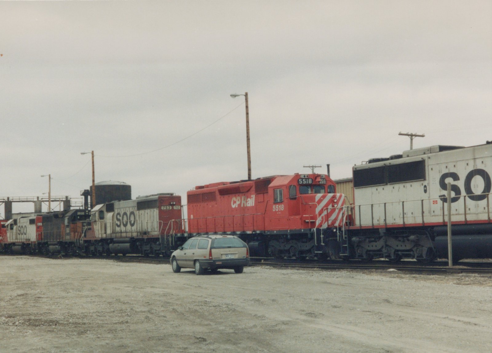 A line of diesel locomotives from CP Rail and the Soo Line are parked in a gravel yard under an overcast sky, with a beige station wagon in the foreground.