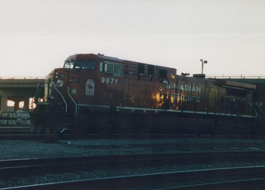 A Canadian Pacific diesel locomotive, number 9671, in maroon paint with white lettering and a special decal, is parked on railroad tracks beneath an overpass during dusk.