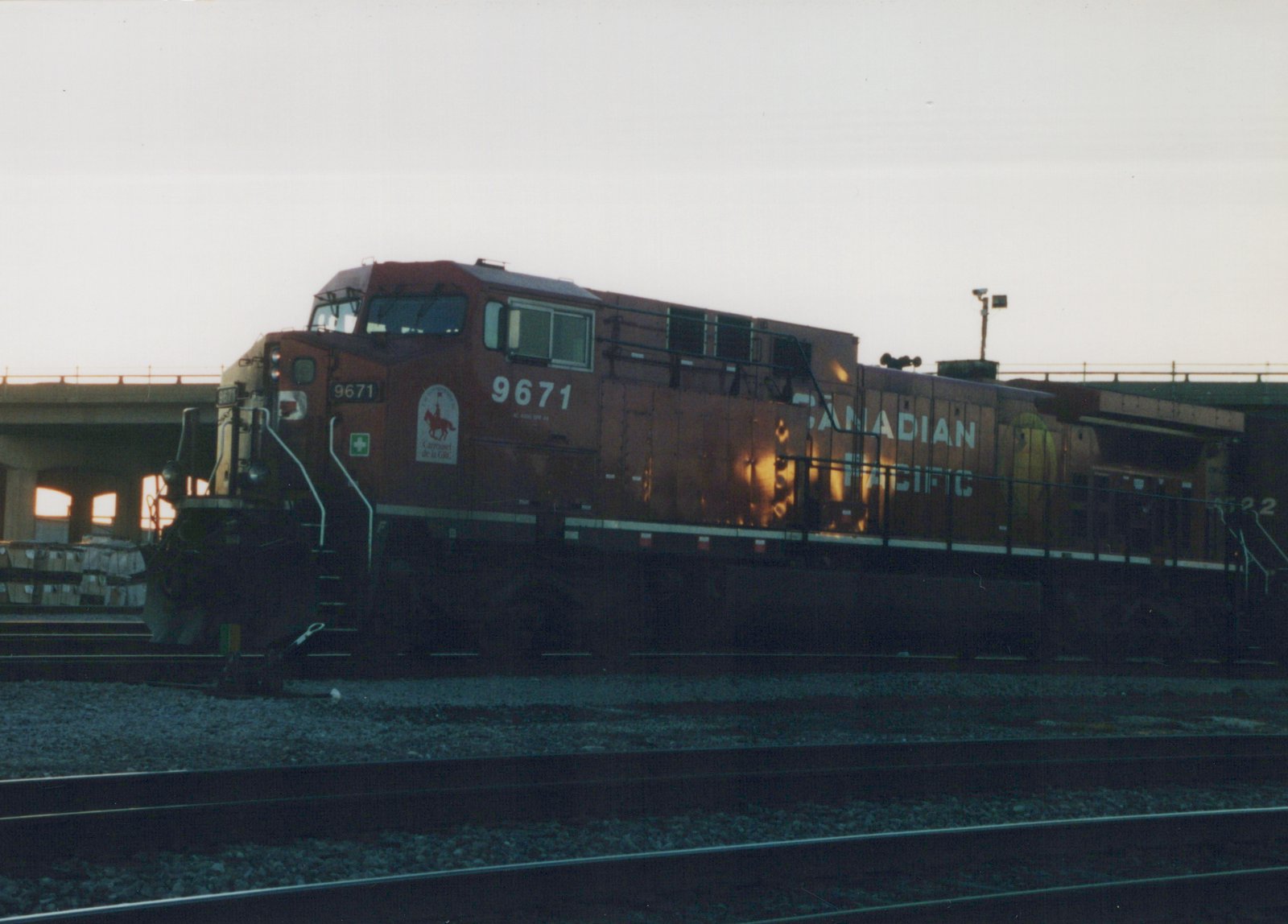 A Canadian Pacific diesel locomotive, number 9671, in maroon paint with white lettering and a special decal, is parked on railroad tracks beneath an overpass during dusk.