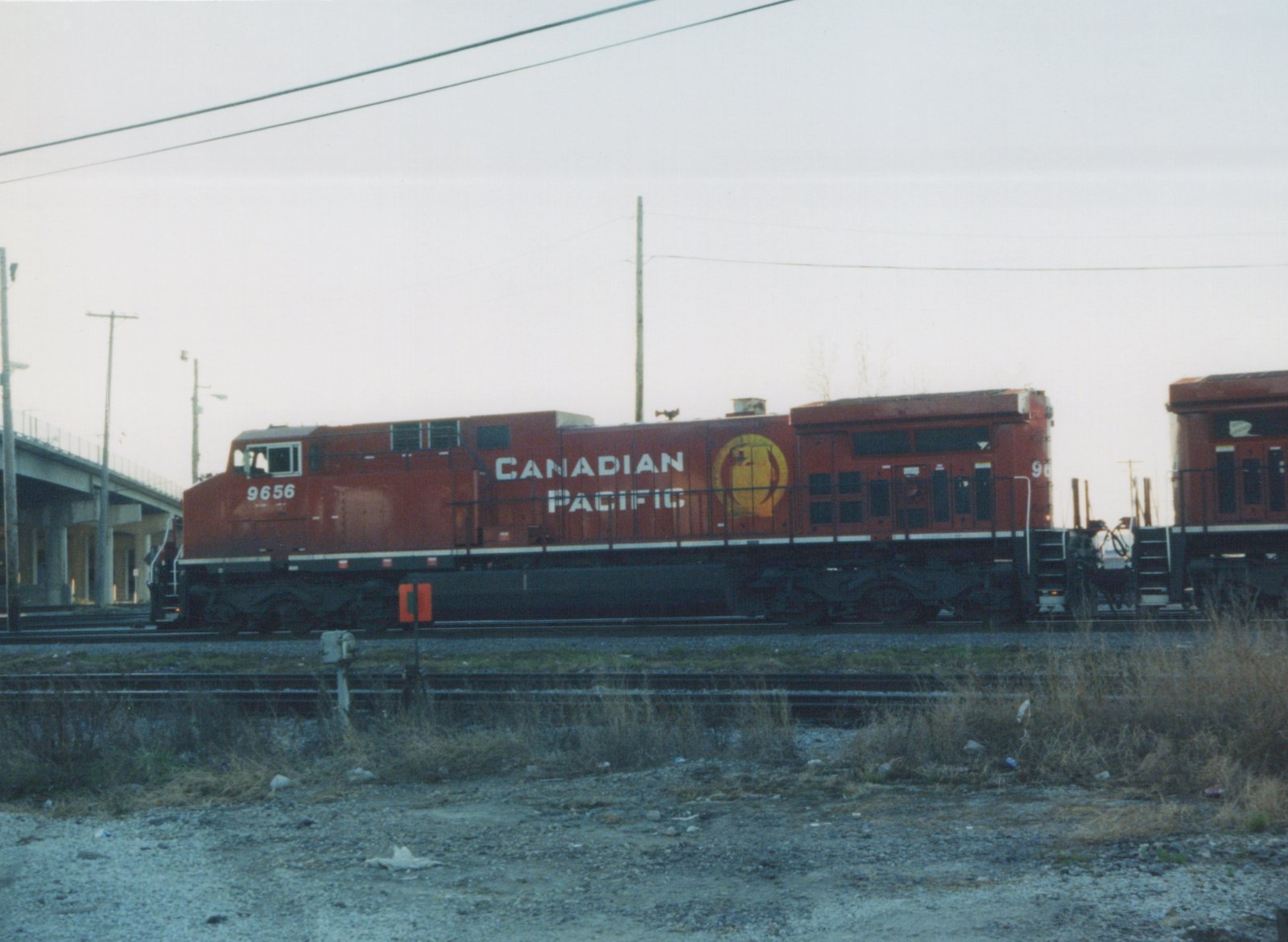 A red Canadian Pacific diesel locomotive numbered 9656 is shown from the side in a rail yard, with a second matching locomotive behind it and a concrete overpass to the left.