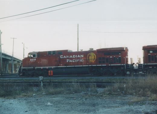 A red Canadian Pacific diesel locomotive numbered 9656 is shown from the side in a rail yard, with a second matching locomotive behind it and a concrete overpass to the left.
