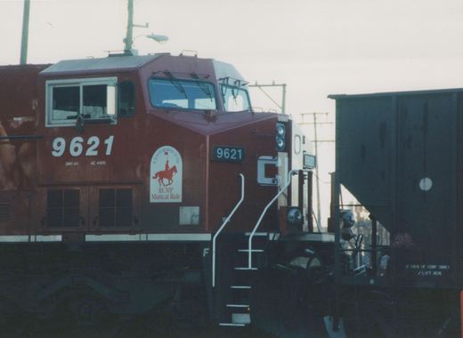 A red Canadian Pacific locomotive numbered 9621 is shown from a three-quarter front view in a railyard, with a dark freight car next to it.