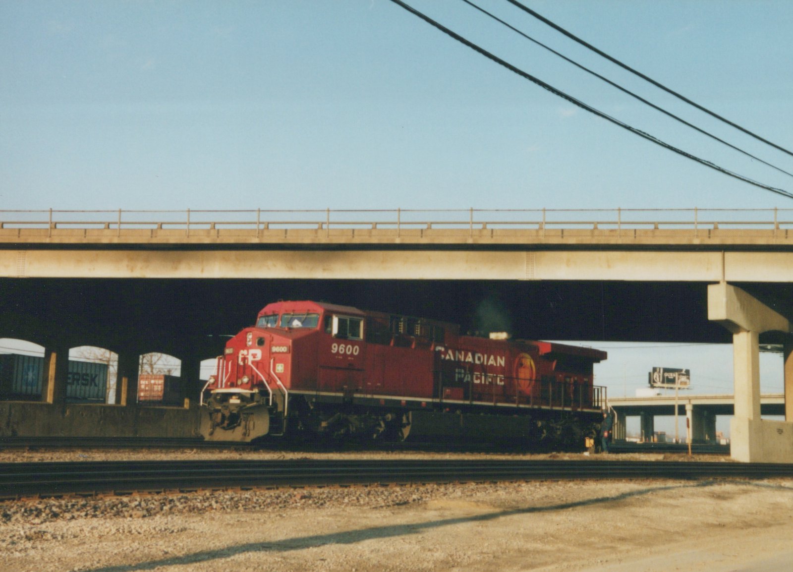 A red Canadian Pacific locomotive number 9600 is captured from a three-quarter front view as it moves under an elevated highway bridge, with clear skies above and a person standing near the tracks.