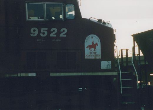 A close-up of a dark-colored Canadian Pacific locomotive numbered 9522, with a decal for the RCMP Musical Ride on its side, set in a yard during dusk with overcast lighting.