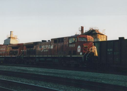A side view of a red CP Rail System locomotive numbered 9522, with a second locomotive and a freight car behind it, on a railroad track in a yard at dusk.