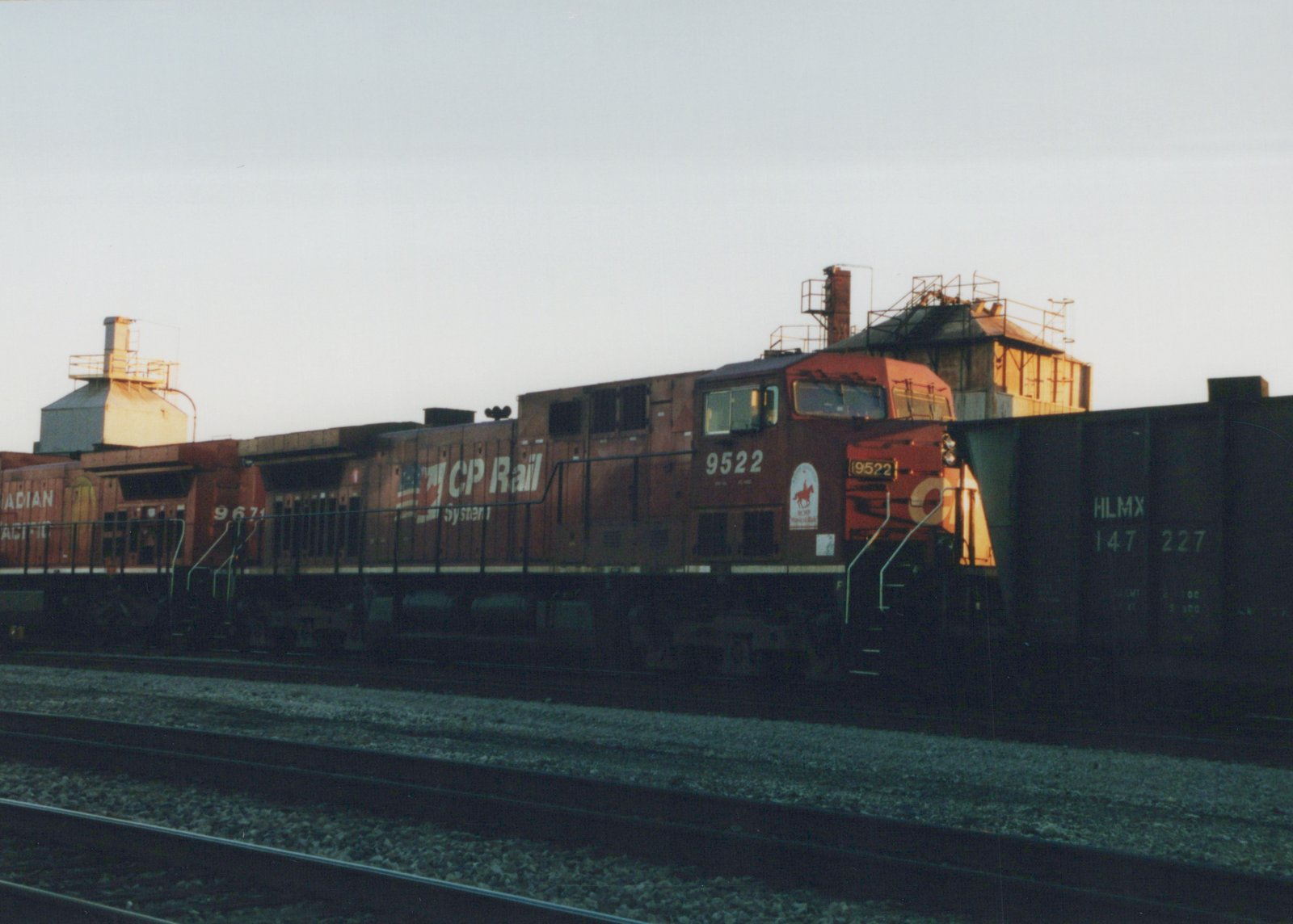 A side view of a red CP Rail System locomotive numbered 9522, with a second locomotive and a freight car behind it, on a railroad track in a yard at dusk.