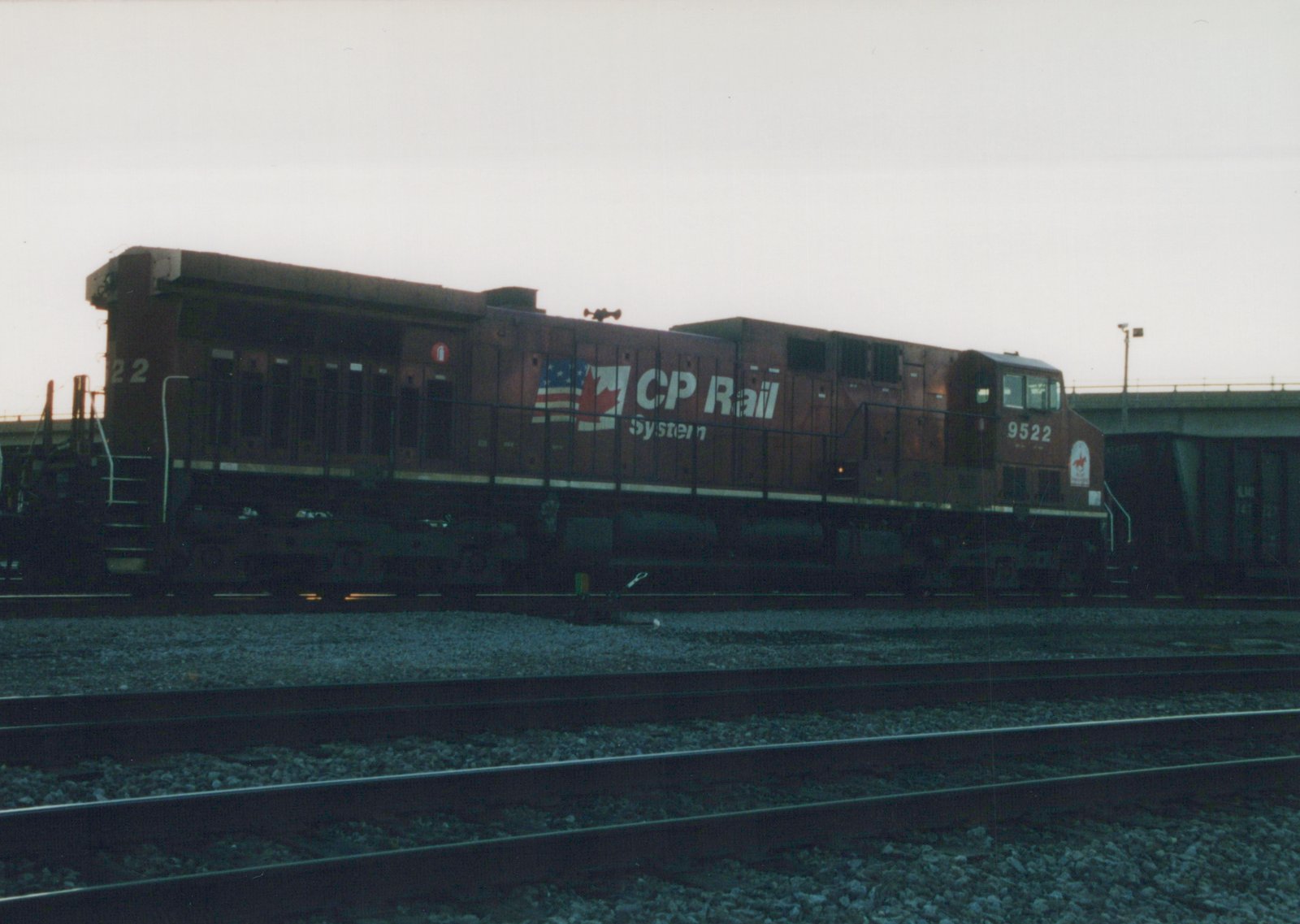 A side view of a maroon CP Rail System diesel locomotive, number 9522, parked on multiple tracks in a yard with a bridge in the background during dusk.