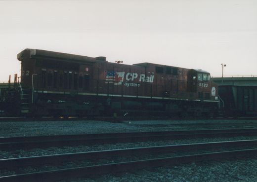 A side view of a maroon CP Rail System diesel locomotive, number 9522, parked on multiple tracks in a yard with a bridge in the background during dusk.