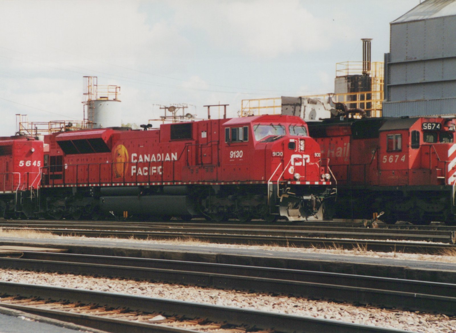 A side view of three red Canadian Pacific GP9 diesel locomotives, numbered 9130, 5645, and 5674, lined up on parallel tracks in a rail yard with industrial buildings in the background.