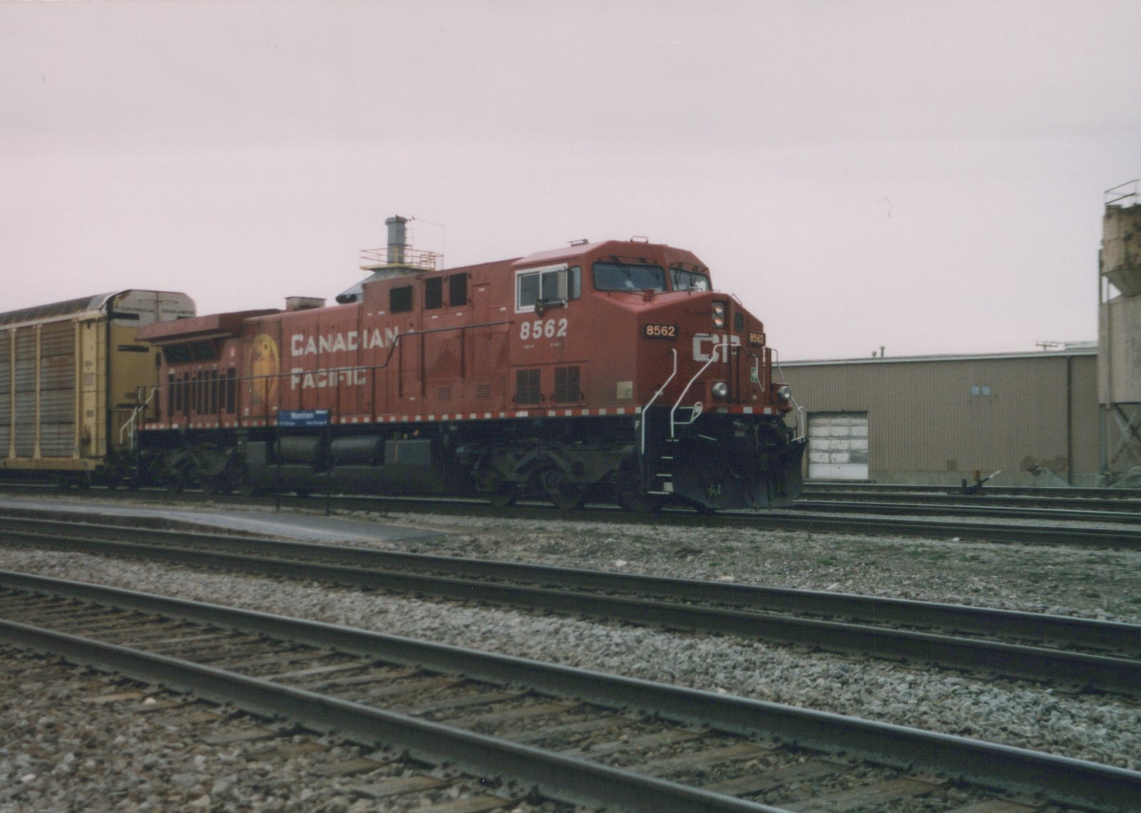 A red Canadian Pacific diesel locomotive, number 8562, is parked on tracks in a rail yard next to a building, with a yellow freight car coupled behind it.