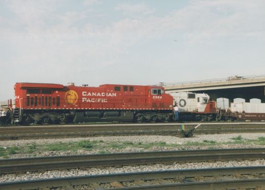 A red Canadian Pacific locomotive numbered 8564 and a white and red Santa Fe locomotive numbered 4445 are parked on parallel tracks in a rail yard with a freight car and an overpass in the background.