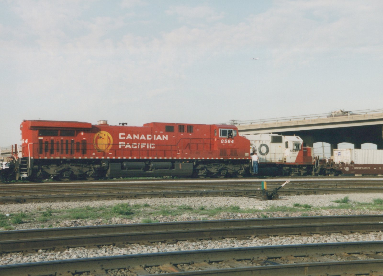 A red Canadian Pacific locomotive numbered 8564 and a white and red Santa Fe locomotive numbered 4445 are parked on parallel tracks in a rail yard with a freight car and an overpass in the background.