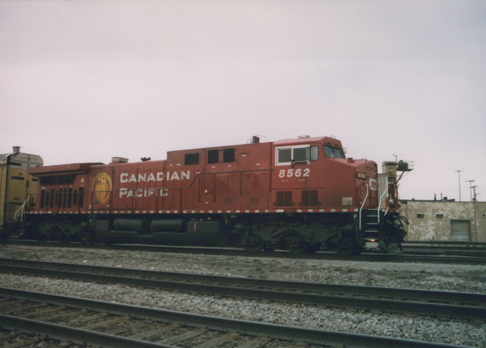 A red Canadian Pacific locomotive numbered 8562 sits on the tracks in a yard, with a yellow freight car behind it and a building in the background under a gray sky.