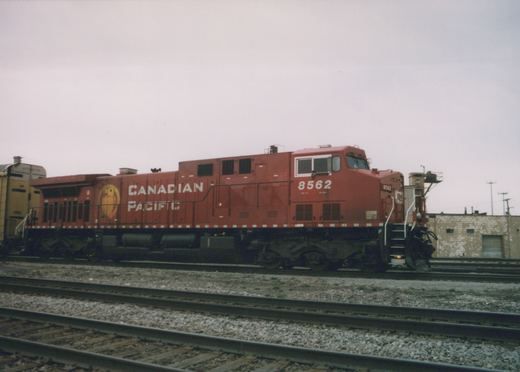 A red Canadian Pacific locomotive numbered 8562 sits on the tracks in a yard, with a yellow freight car behind it and a building in the background under a gray sky.