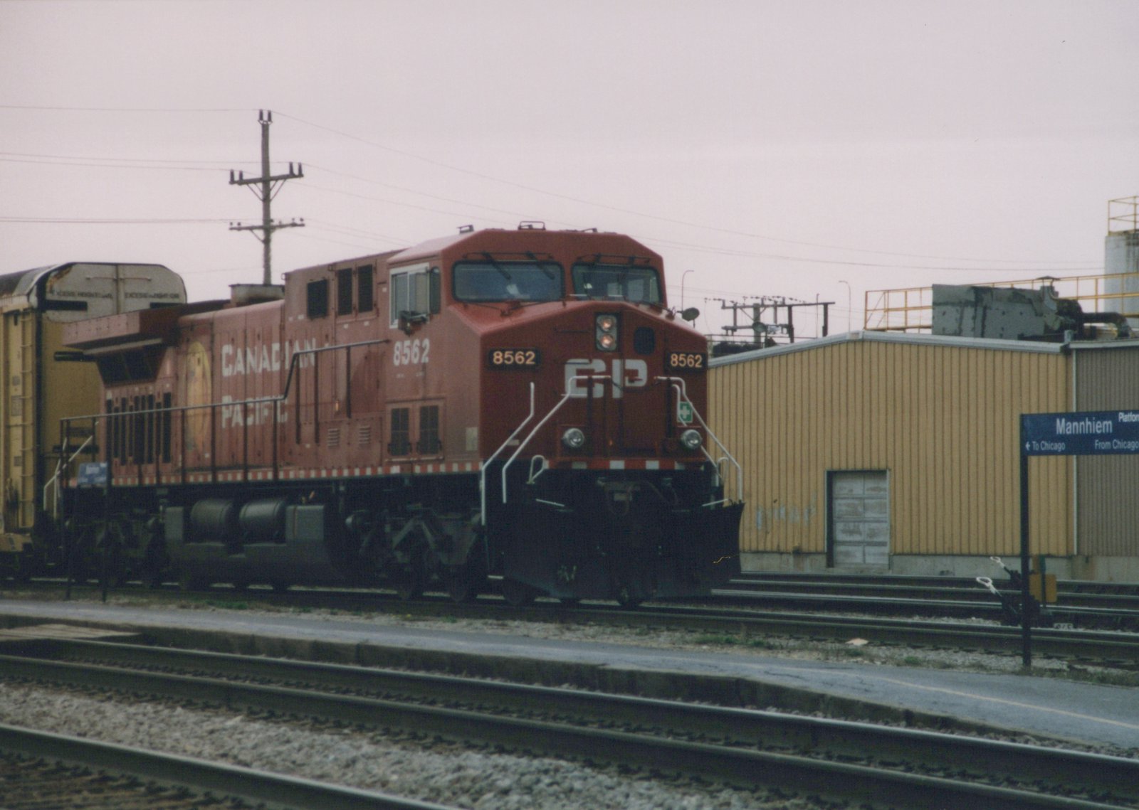 A red Canadian Pacific diesel locomotive numbered 8562 sits on a railroad track next to a blue station sign for Mannhiem, with a yellow freight car behind it.