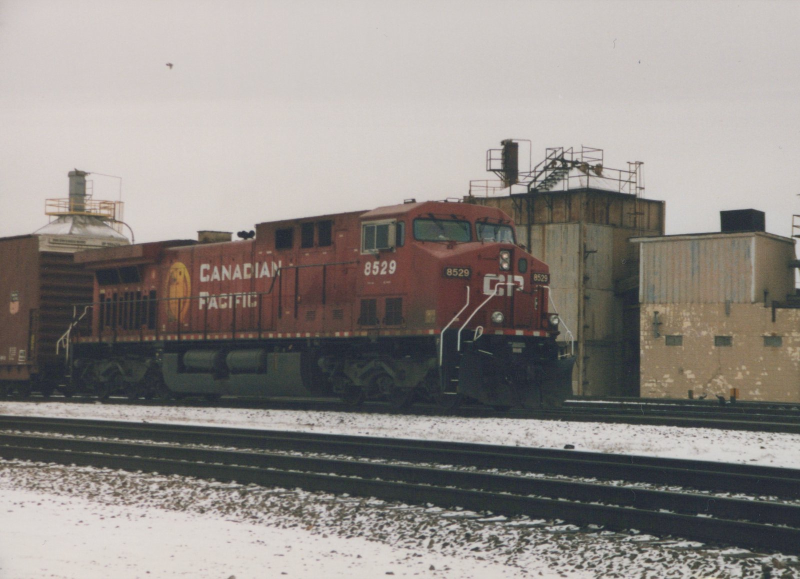 A red Canadian Pacific diesel locomotive, number 8529, sits on railroad tracks in a snowy yard with industrial buildings in the background.