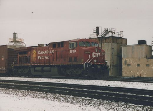 A red Canadian Pacific diesel locomotive, number 8529, sits on railroad tracks in a snowy yard with industrial buildings in the background.