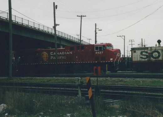 A red Canadian Pacific diesel locomotive numbered 8527 travels on a railroad track under a highway overpass, with a freight car marked 'SO' visible behind it.
