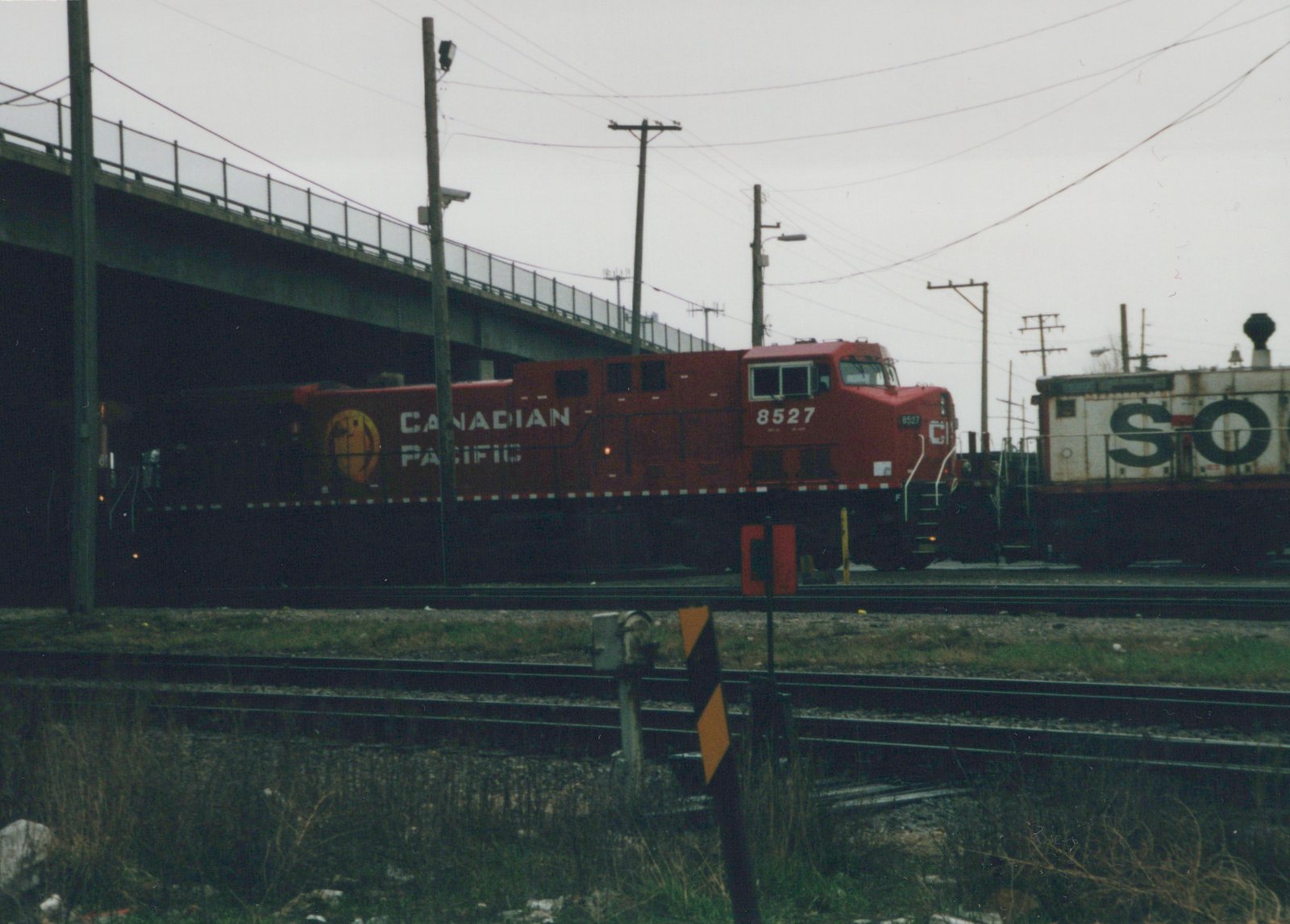 A red Canadian Pacific diesel locomotive numbered 8527 travels on a railroad track under a highway overpass, with a freight car marked 'SO' visible behind it.
