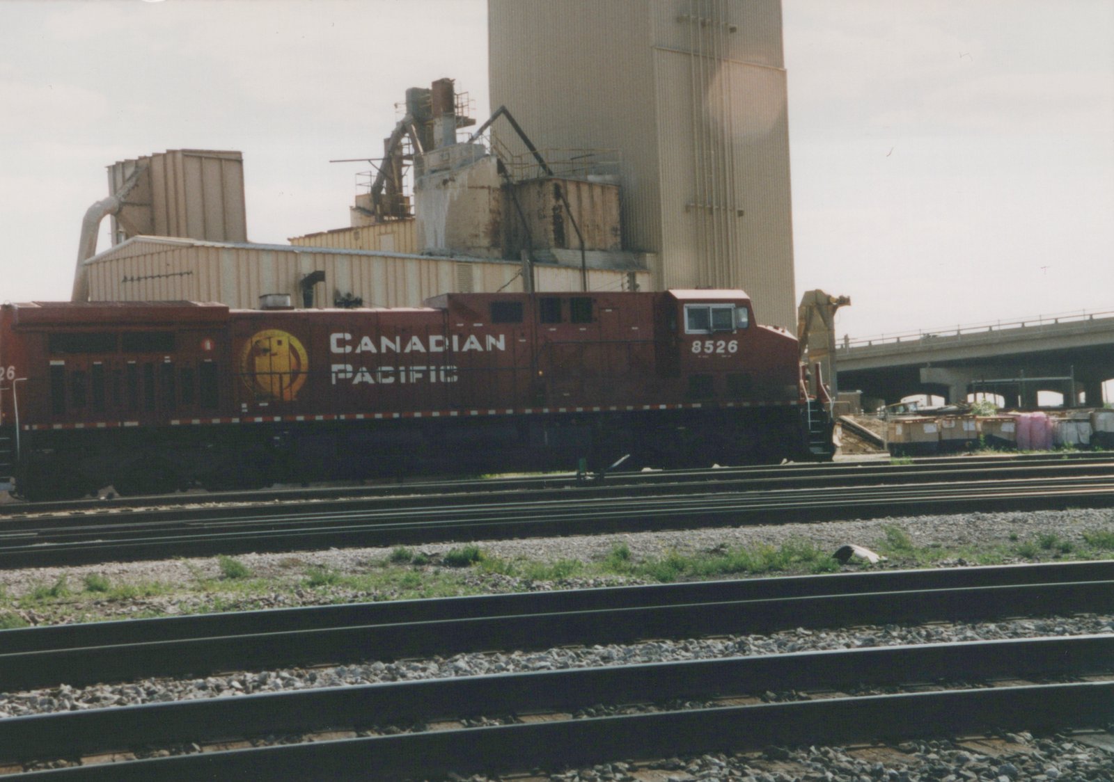 A maroon Canadian Pacific diesel locomotive with the number 8526 on its cab side sits on multiple train tracks in front of a large industrial building and a highway overpass under an overcast sky.
