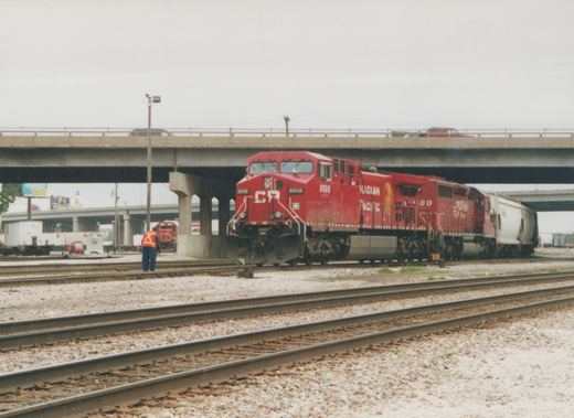 A red Canadian Pacific locomotive, number 8508, sits on railroad tracks in a yard under a bridge, with a worker nearby and another locomotive in the background.