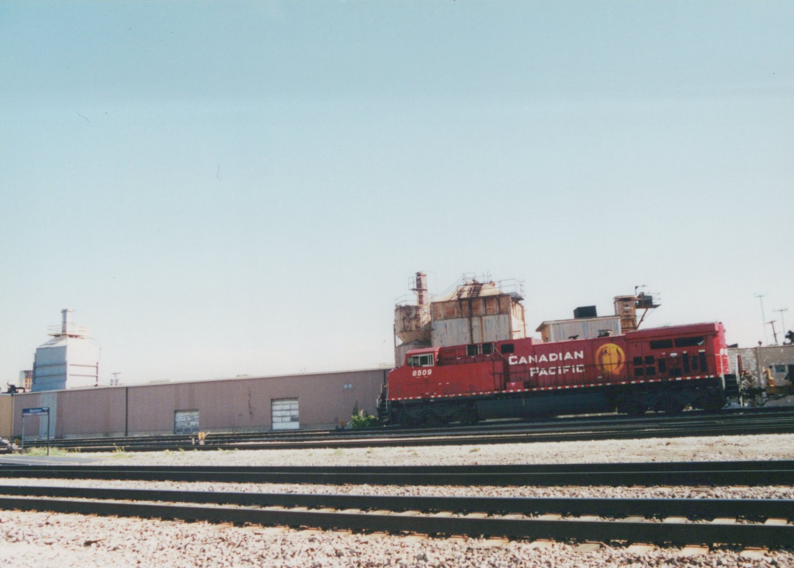 A red Canadian Pacific diesel locomotive numbered 8509 sits on tracks in a rail yard with industrial buildings in the background under a clear blue sky.