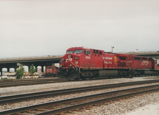 A red Canadian Pacific freight train with locomotive 8508 leading, followed by locomotive 5680, on tracks under a concrete overpass on a cloudy day.