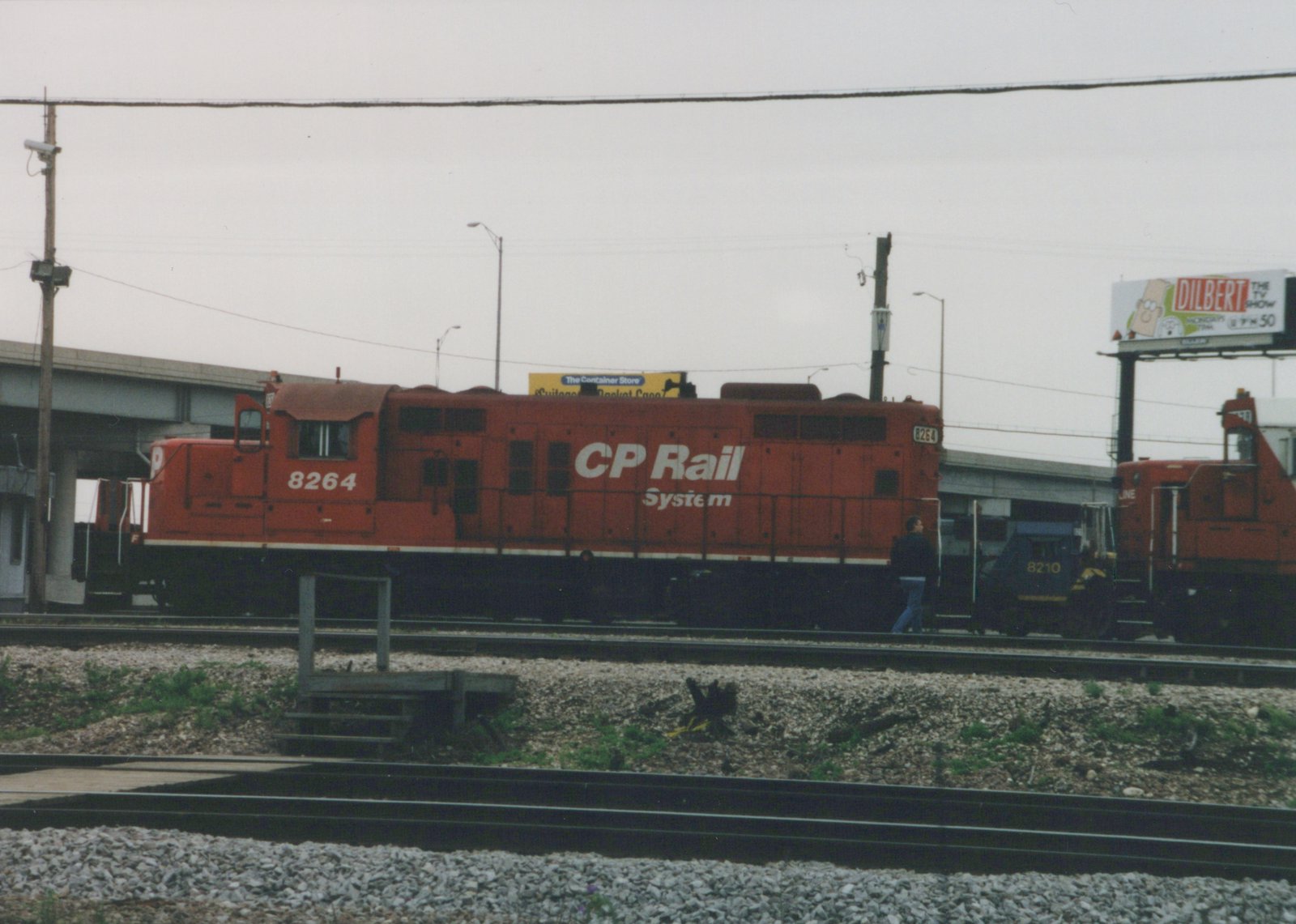 A red CP Rail System locomotive numbered 8264, alongside track infrastructure and a billboard advertising the TV show Dilbert.