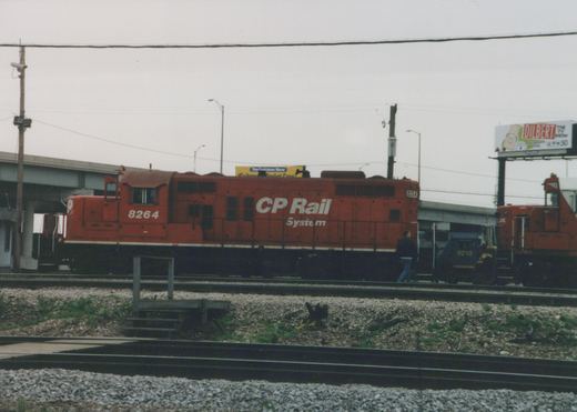 A red CP Rail System locomotive numbered 8264, alongside track infrastructure and a billboard advertising the TV show Dilbert.