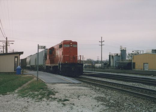 A red diesel locomotive with the number 8263 on its front is stopped at a platform in a rail yard, with multiple tracks and industrial buildings in the background.