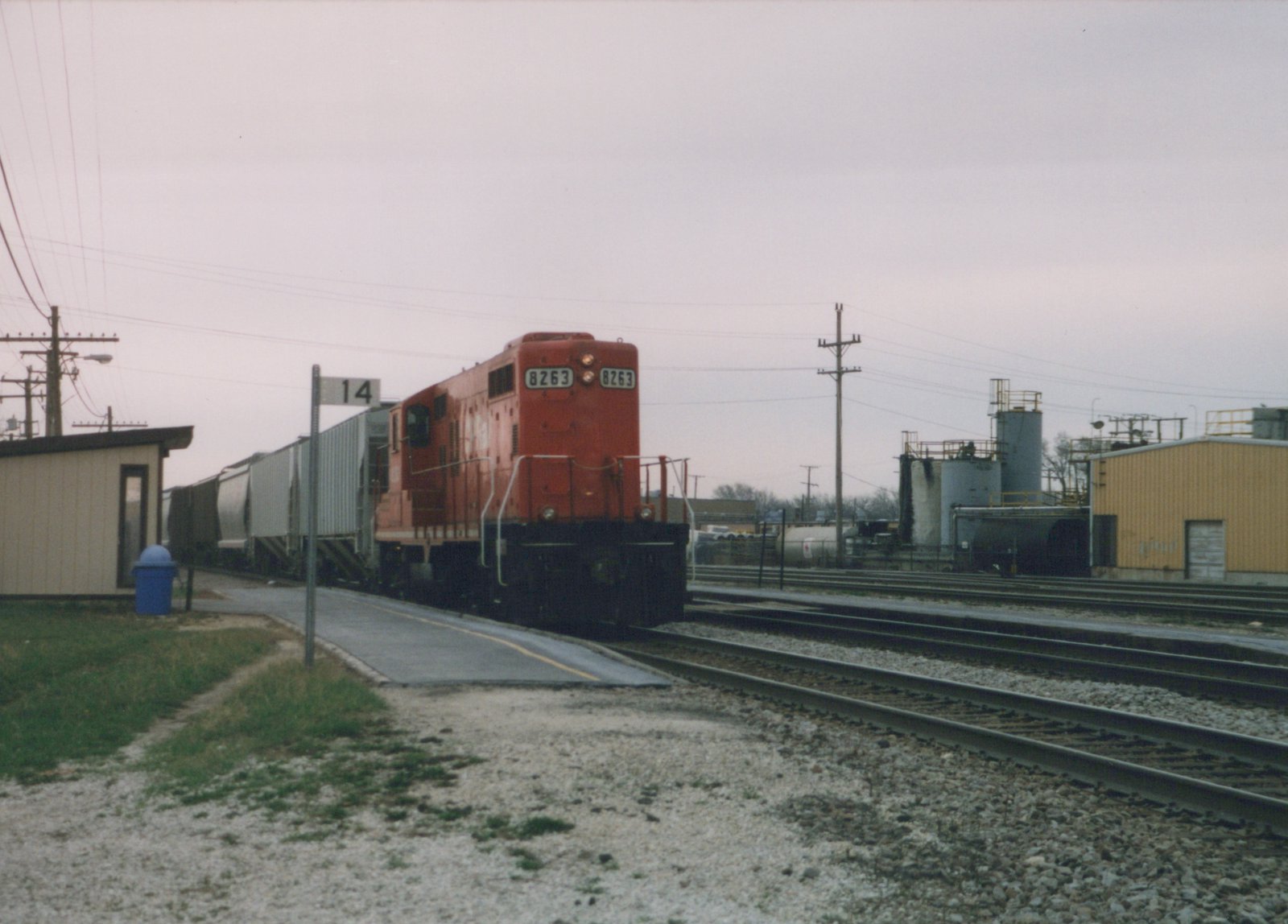 A red diesel locomotive with the number 8263 on its front is stopped at a platform in a rail yard, with multiple tracks and industrial buildings in the background.