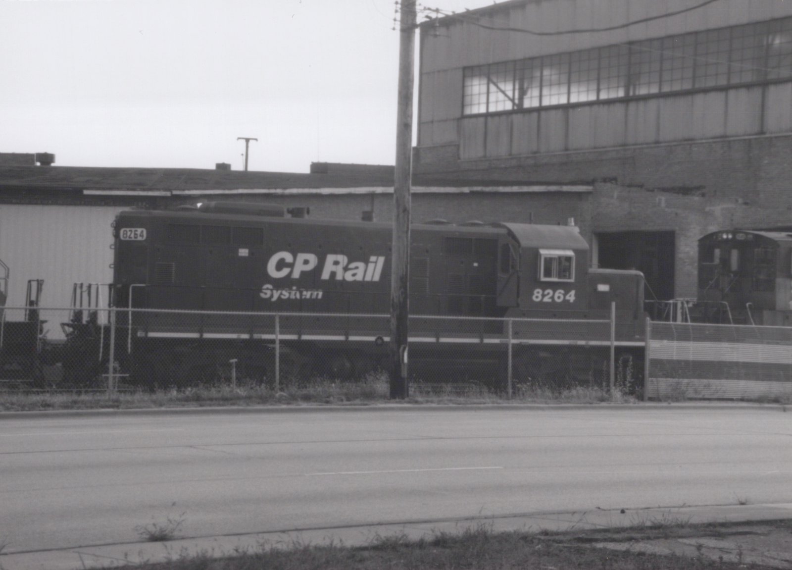 A black-and-white photo of a CP Rail System diesel locomotive numbered 8264, parked behind a chain-link fence next to an industrial building.