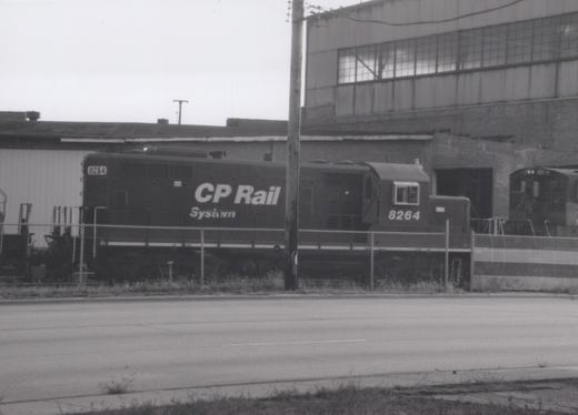 A black-and-white photo of a CP Rail System diesel locomotive numbered 8264, parked behind a chain-link fence next to an industrial building.