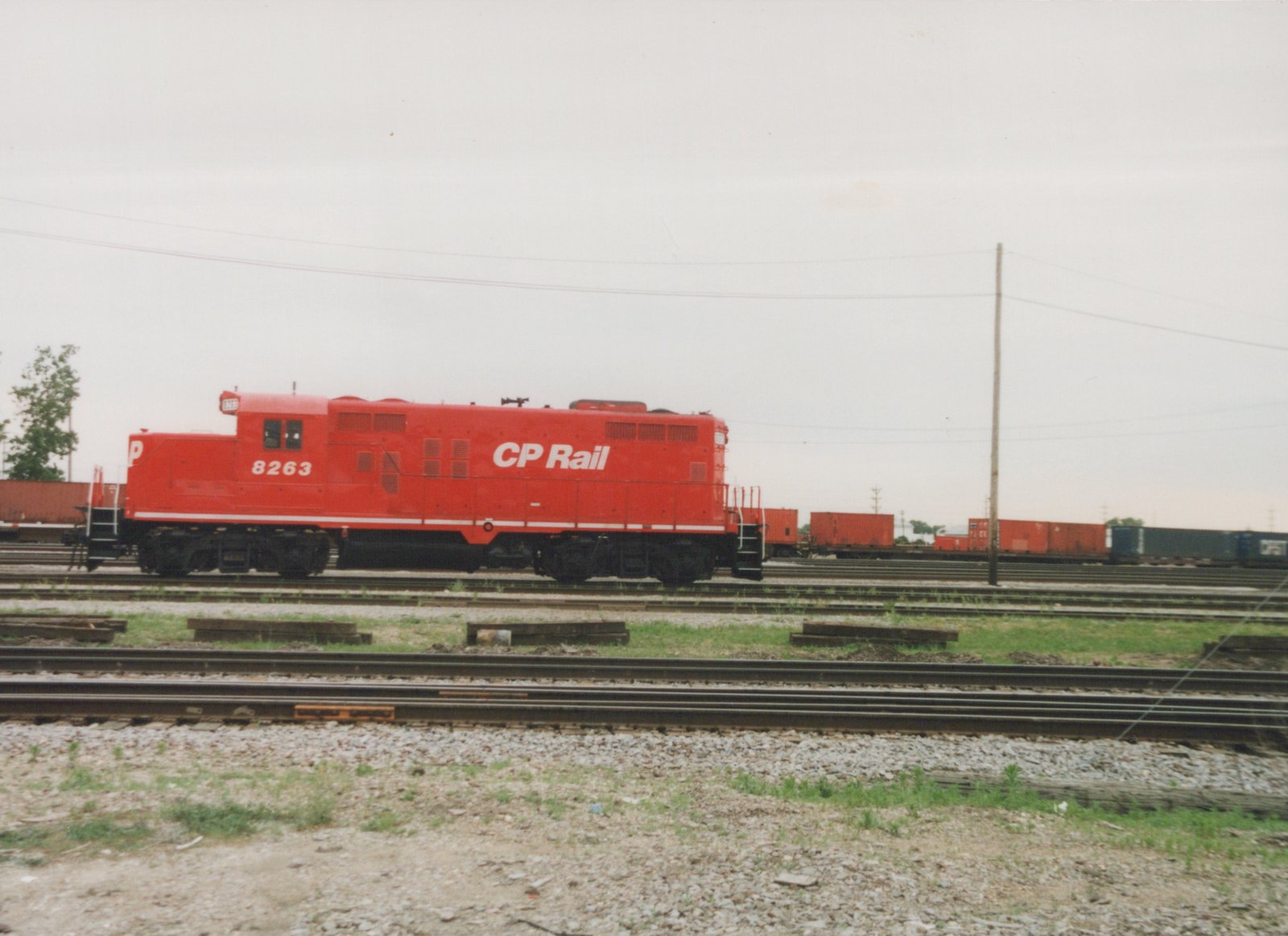 A red Canadian Pacific locomotive numbered 8263 is parked on a set of tracks in a large rail yard, with several freight cars and containers in the background under an overcast sky.