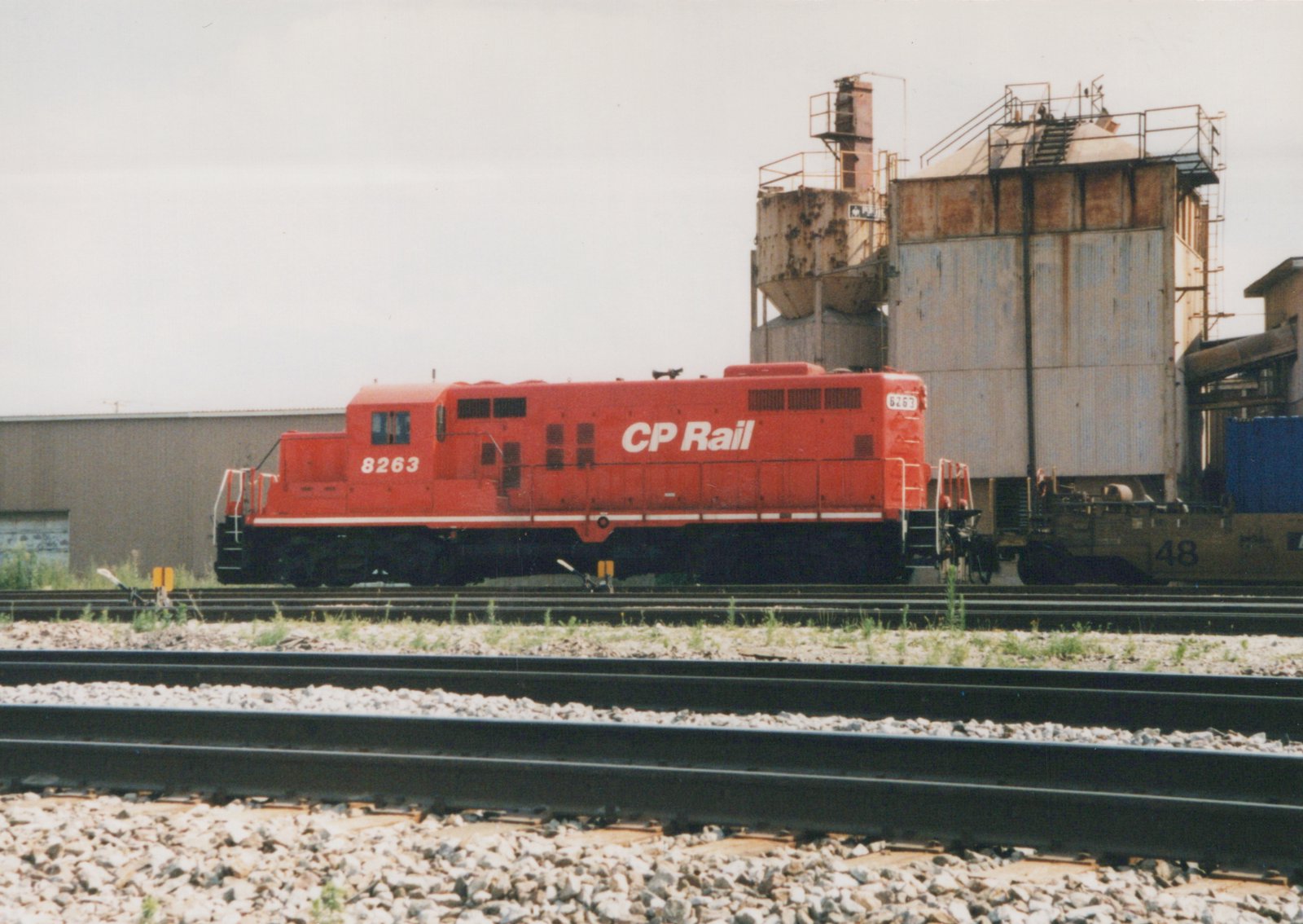A side view of a red CP Rail diesel locomotive, number 8263, parked on railroad tracks next to industrial buildings under an overcast sky.