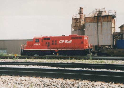 A side view of a red CP Rail diesel locomotive, number 8263, parked on railroad tracks next to industrial buildings under an overcast sky.