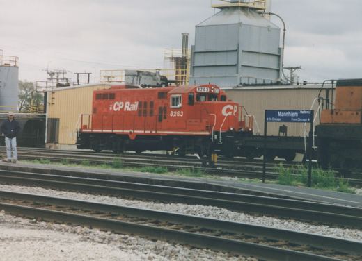A red CP Rail System locomotive numbered 8263 sits on tracks at a station platform with a blue sign reading 'Mannhiem Platform 2', under a cloudy sky.