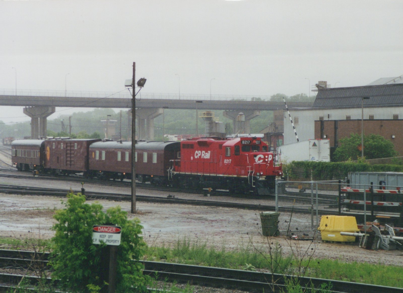 A red CP Rail diesel locomotive, number 8217, is shown pulling three maroon passenger cars on a set of tracks in a rail yard. A concrete overpass is visible in the background, and a 'DANGER KEEP OFF C.P. RAIL PROPERTY' sign stands in the foreground.