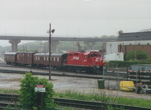 A red CP Rail diesel locomotive, number 8217, is shown pulling three maroon passenger cars on a set of tracks in a rail yard. A concrete overpass is visible in the background, and a 'DANGER KEEP OFF C.P. RAIL PROPERTY' sign stands in the foreground.